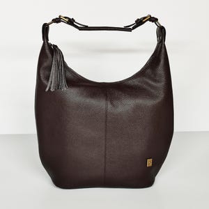 May include: A dark brown hobo bag with a pebbled texture, featuring a single shoulder strap with brass-toned hardware, a decorative gray leather tassel attached to the side, and a small rectangular gold-toned logo plaque on the lower right corner.