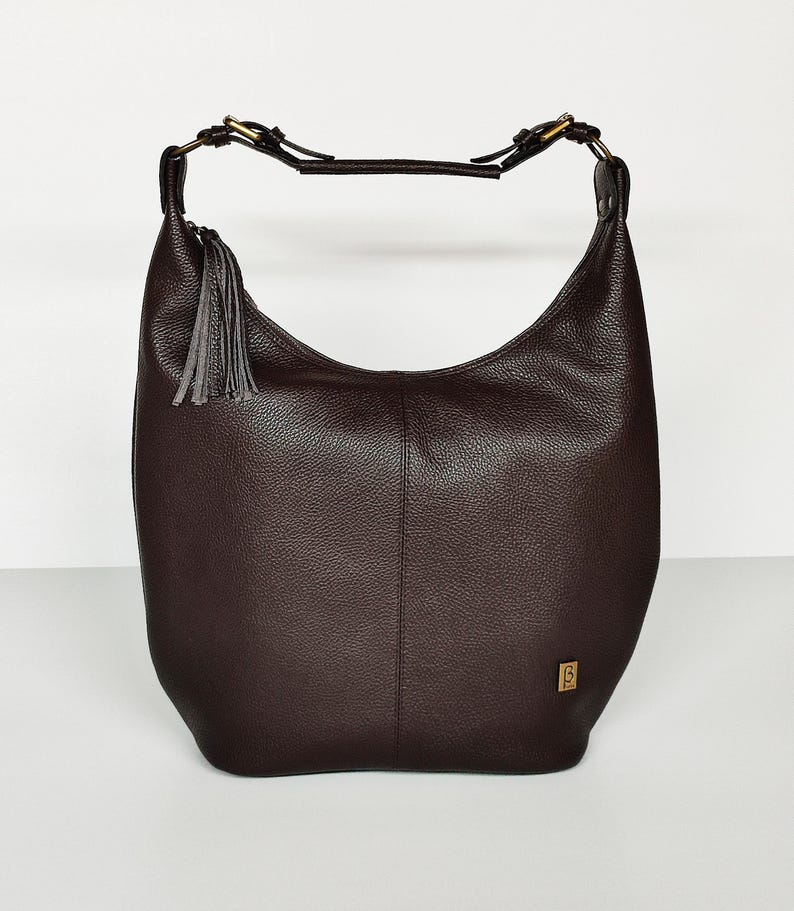 May include: A dark brown hobo bag with a pebbled texture, featuring a single shoulder strap with brass-toned hardware, a decorative gray leather tassel attached to the side, and a small rectangular gold-toned logo plaque on the lower right corner.