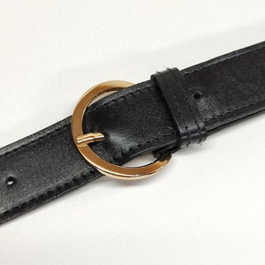 May include: A black belt featuring a smooth texture and a polished gold-tone circular buckle. The belt includes a matching black leather-like keeper loop and visible stitching along the edges of the strap, with pre-punched holes for adjustment.
