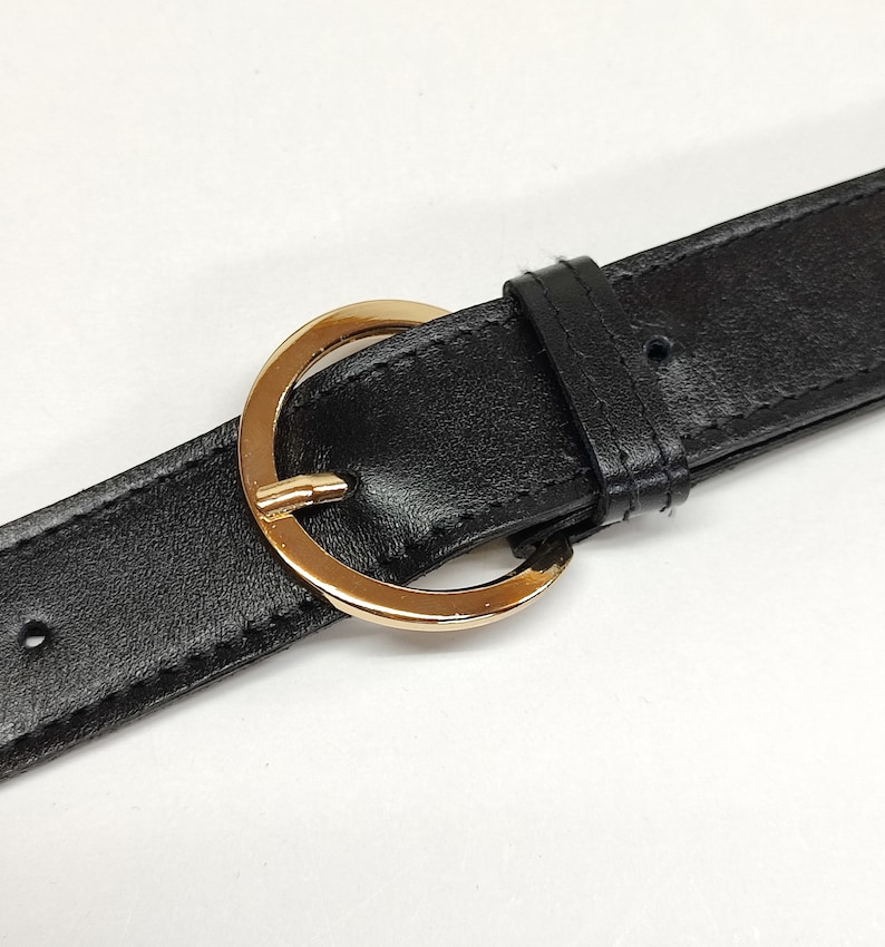 May include: A black belt featuring a smooth texture and a polished gold-tone circular buckle. The belt includes a matching black leather-like keeper loop and visible stitching along the edges of the strap, with pre-punched holes for adjustment.