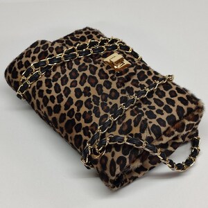 May include: A rectangular shoulder bag featuring a brown and black leopard print pattern with a textured fur-like finish. The bag includes a gold-tone metal turn-lock closure and a chain strap interwoven with black material for a secure carry.