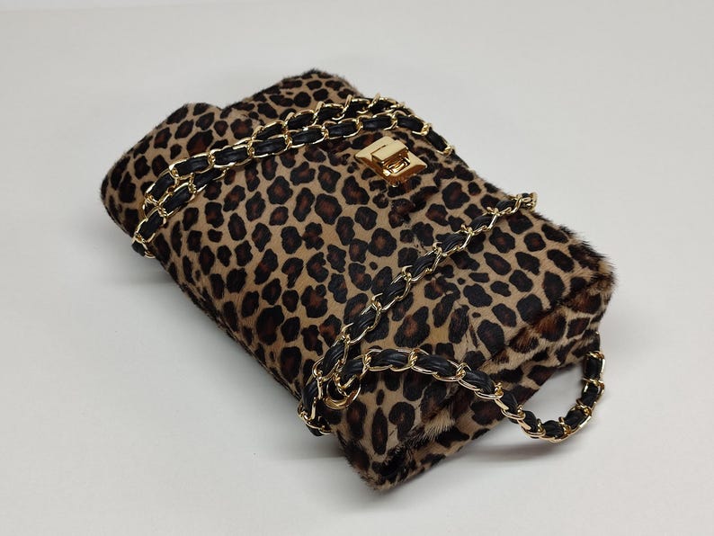May include: A rectangular shoulder bag featuring a brown and black leopard print pattern with a textured fur-like finish. The bag includes a gold-tone metal turn-lock closure and a chain strap interwoven with black material for a secure carry.