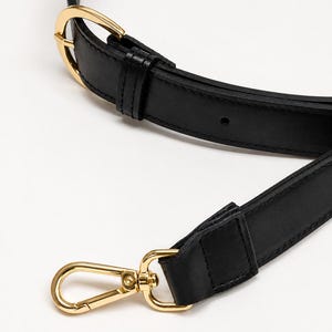 May include: A black leather bag strap featuring gold-toned metal hardware, including an adjustable buckle with a prong and a swivel lobster clasp at the end. The strap has stitched edges and a smooth finish, designed for attaching to a handbag.
