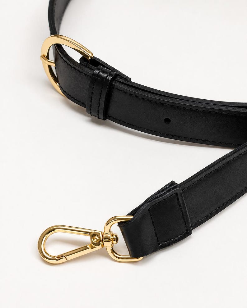 May include: A black leather bag strap featuring gold-toned metal hardware, including an adjustable buckle with a prong and a swivel lobster clasp at the end. The strap has stitched edges and a smooth finish, designed for attaching to a handbag.