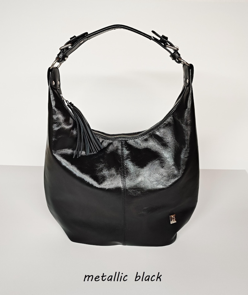 May include: A metallic black hobo bag featuring a single shoulder strap with silver-toned buckles, a top zipper closure, and a decorative leather tassel. A small metallic logo plaque is attached to the lower right side of the bag.