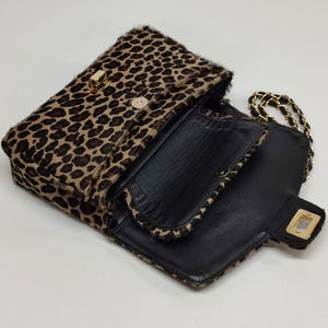May include: A leopard print shoulder bag with a textured fur-like exterior, featuring a black interior lining, a gold-tone rectangular turn-lock closure on the front flap, and a gold-tone chain strap interwoven with black material.