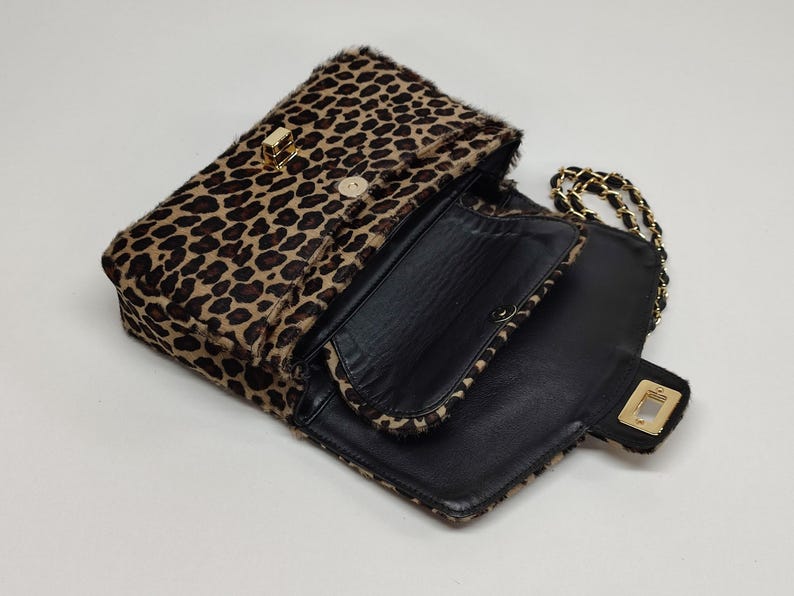 May include: A leopard print shoulder bag with a textured fur-like exterior, featuring a black interior lining, a gold-tone rectangular turn-lock closure on the front flap, and a gold-tone chain strap interwoven with black material.