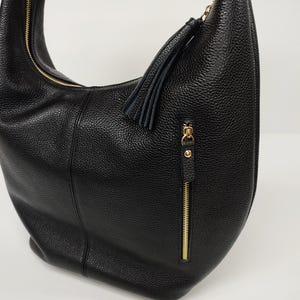 May include: A black hobo bag with a pebbled texture, featuring a gold-tone zipper closure along the top and a vertical gold-tone zippered pocket on the front. A black leather tassel is attached to the zipper pull of the front pocket.