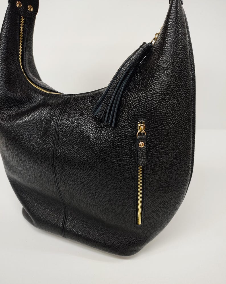 May include: A black hobo bag with a pebbled texture, featuring a gold-tone zipper closure along the top and a vertical gold-tone zippered pocket on the front. A black leather tassel is attached to the zipper pull of the front pocket.