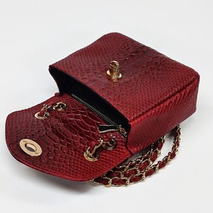 May include: A red, textured handbag with a gold chain strap and gold hardware. The bag is open, revealing a black interior and a small zippered pocket. The exterior has a snakeskin pattern.