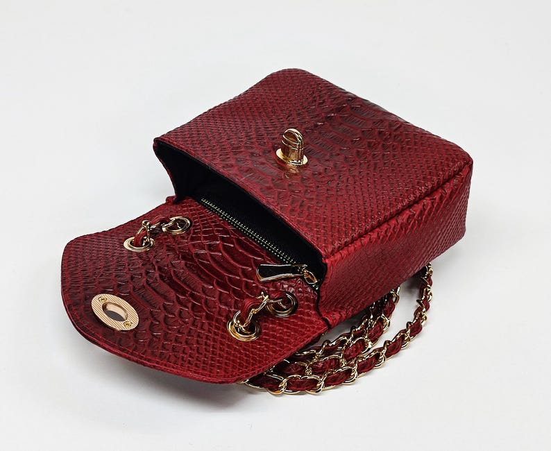 May include: A red, textured handbag with a gold chain strap and gold hardware. The bag is open, revealing a black interior and a small zippered pocket. The exterior has a snakeskin pattern.