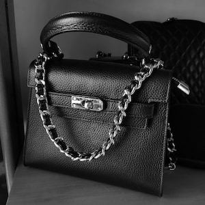 Puede incluir: Un bolso de mano de cuero negro con una correa de cadena plateada y un cierre de candado plateado.