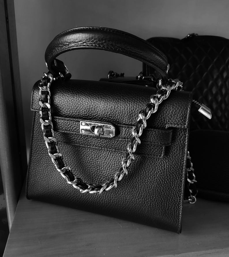 Puede incluir: Un bolso de mano de cuero negro con una correa de cadena plateada y un cierre de candado plateado.