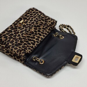 May include: A rectangular shoulder bag featuring a textured leopard print pattern in shades of brown and black. The bag has a gold-tone metal turn-lock closure on the front flap and a gold-tone chain strap interwoven with black material.
