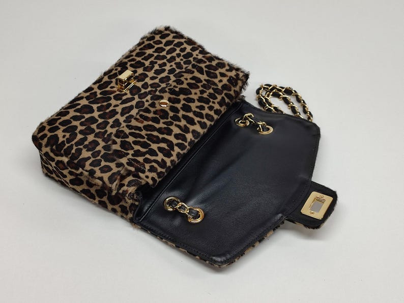 May include: A rectangular shoulder bag featuring a textured leopard print pattern in shades of brown and black. The bag has a gold-tone metal turn-lock closure on the front flap and a gold-tone chain strap interwoven with black material.
