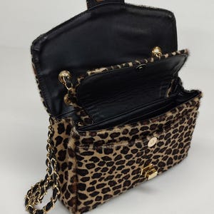 May include: A leopard print shoulder bag with a textured fur-like finish, featuring a gold-tone metal turn-lock closure, a black interior lining, and a gold chain strap interwoven with black material. The bag is shown in an open position.