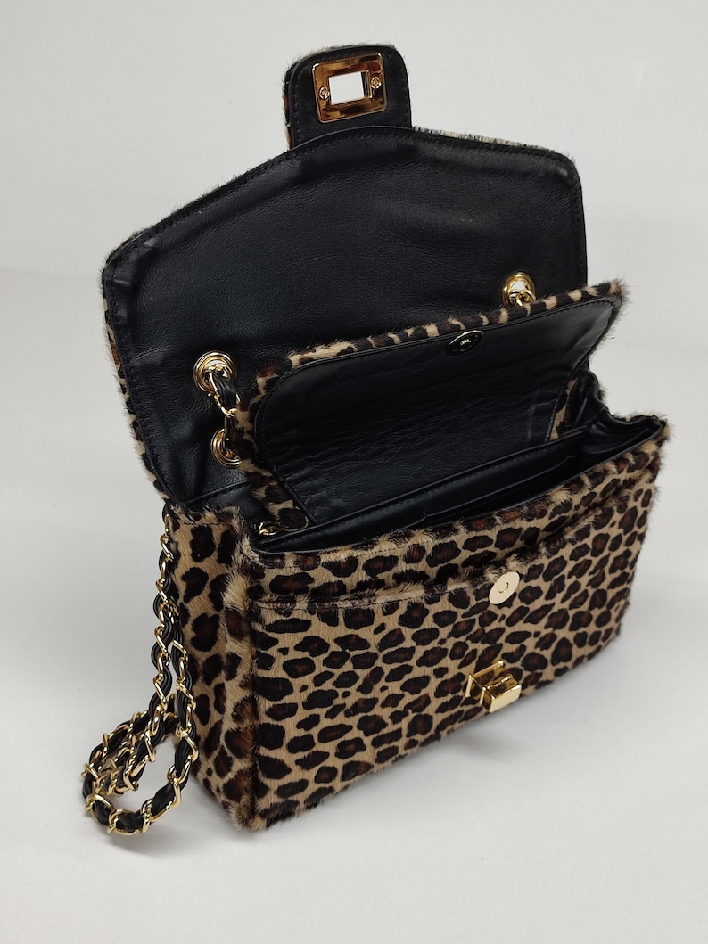 May include: A leopard print shoulder bag with a textured fur-like finish, featuring a gold-tone metal turn-lock closure, a black interior lining, and a gold chain strap interwoven with black material. The bag is shown in an open position.