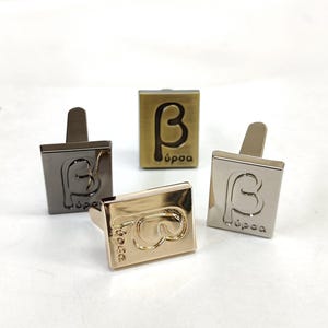 May include: Four rectangular metal cufflinks in different finishes including gunmetal, brushed brass, polished gold, and silver. Each cufflink features an embossed Greek beta symbol and the text "bursa" in a lowercase sans-serif font on the front face.