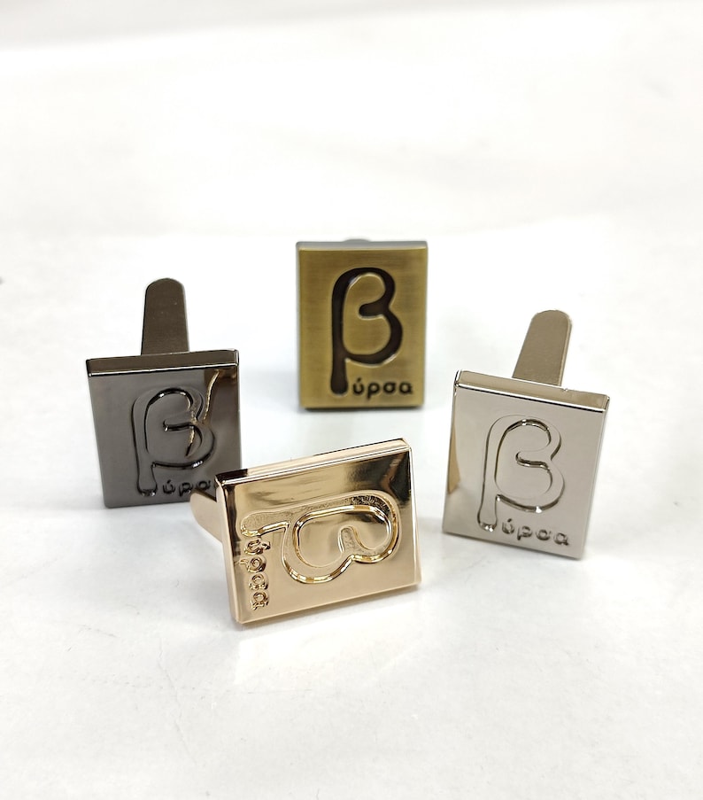 May include: Four rectangular metal cufflinks in different finishes including gunmetal, brushed brass, polished gold, and silver. Each cufflink features an embossed Greek beta symbol and the text "bursa" in a lowercase sans-serif font on the front face.