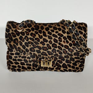 May include: A rectangular shoulder bag featuring a brown and black leopard print pattern with a textured fur-like finish. The bag includes a gold-tone metal turn-lock closure and a chain strap interwoven with black material, attached by gold rings.