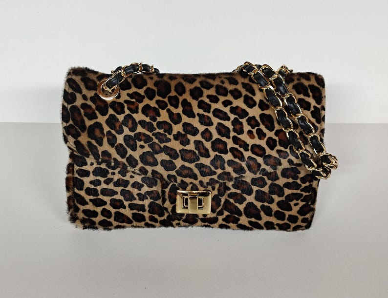 May include: A rectangular shoulder bag featuring a brown and black leopard print pattern with a textured fur-like finish. The bag includes a gold-tone metal turn-lock closure and a chain strap interwoven with black material, attached by gold rings.