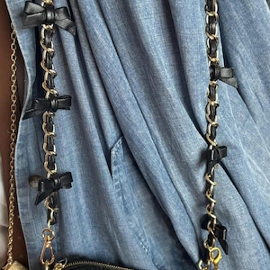 K&ouml;nnte beinhalten: Schwarze und goldene Kettentasche mit schwarzen Schleifenakzenten. Die Tasche hat ein schwarz-wei&szlig;es Karomuster und einen goldenen Rei&szlig;verschluss. Der Kettenriemen ist mit schwarzen Schleifen verziert. Die Tasche h&auml;ngt an einer blauen Jeansjacke.
