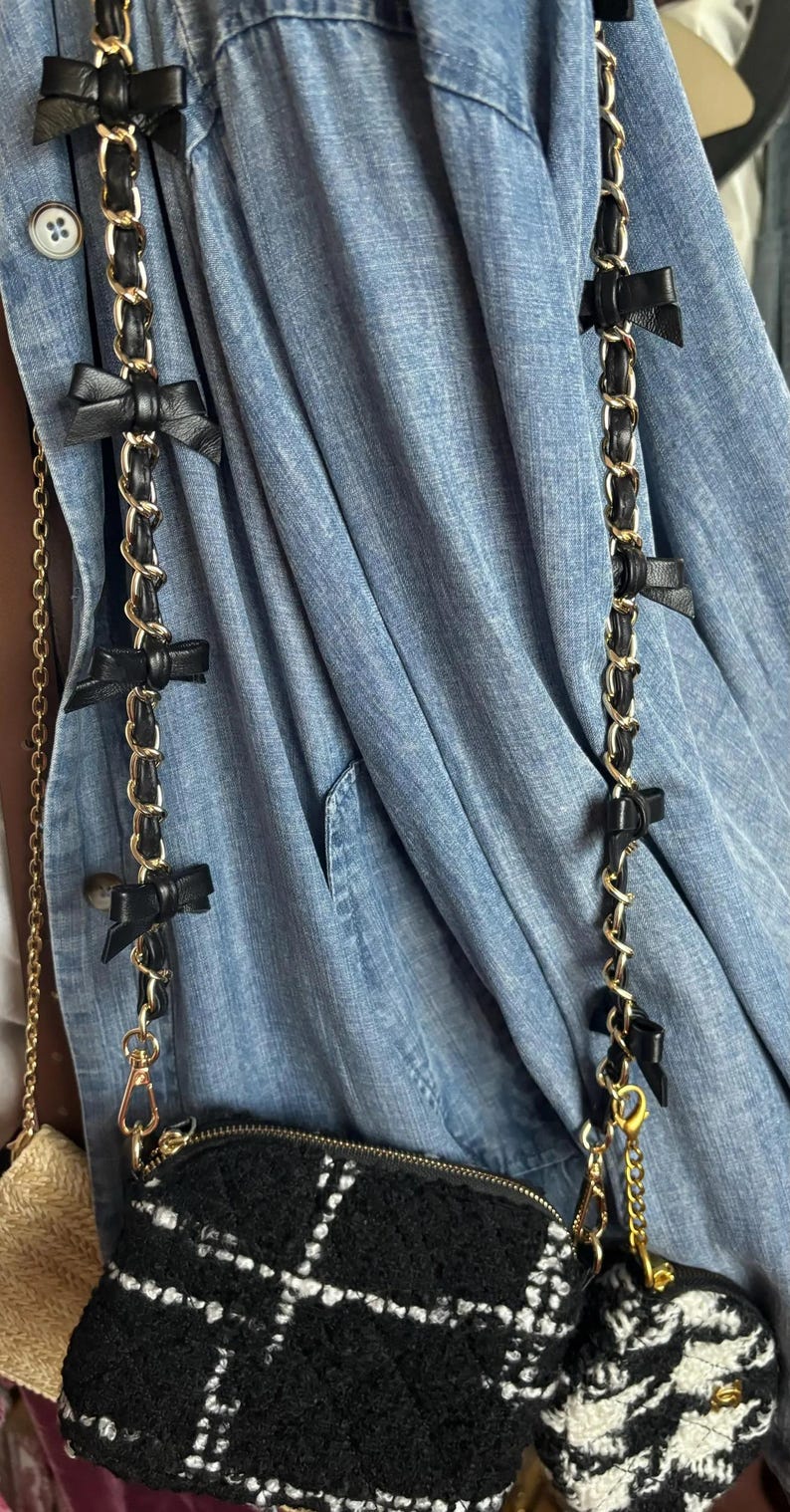 K&ouml;nnte beinhalten: Schwarze und goldene Kettentasche mit schwarzen Schleifenakzenten. Die Tasche hat ein schwarz-wei&szlig;es Karomuster und einen goldenen Rei&szlig;verschluss. Der Kettenriemen ist mit schwarzen Schleifen verziert. Die Tasche h&auml;ngt an einer blauen Jeansjacke.