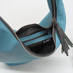May include: A crescent-shaped light blue hobo bag with a pebbled texture, featuring a silver-toned metal zipper closure, a black leather tassel pull, and a black fabric interior lining with a zippered pocket and a label reading Versabags made in Greece.