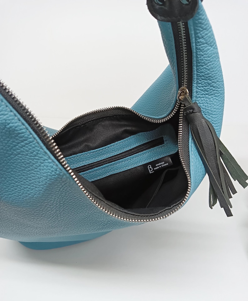 May include: A crescent-shaped light blue hobo bag with a pebbled texture, featuring a silver-toned metal zipper closure, a black leather tassel pull, and a black fabric interior lining with a zippered pocket and a label reading Versabags made in Greece.