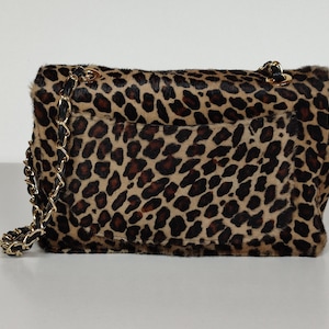 May include: A rectangular shoulder bag featuring a textured leopard print pattern in shades of tan, brown, and black. The bag includes a gold-tone metal chain strap interwoven with black material and a flap closure with a visible exterior pocket.