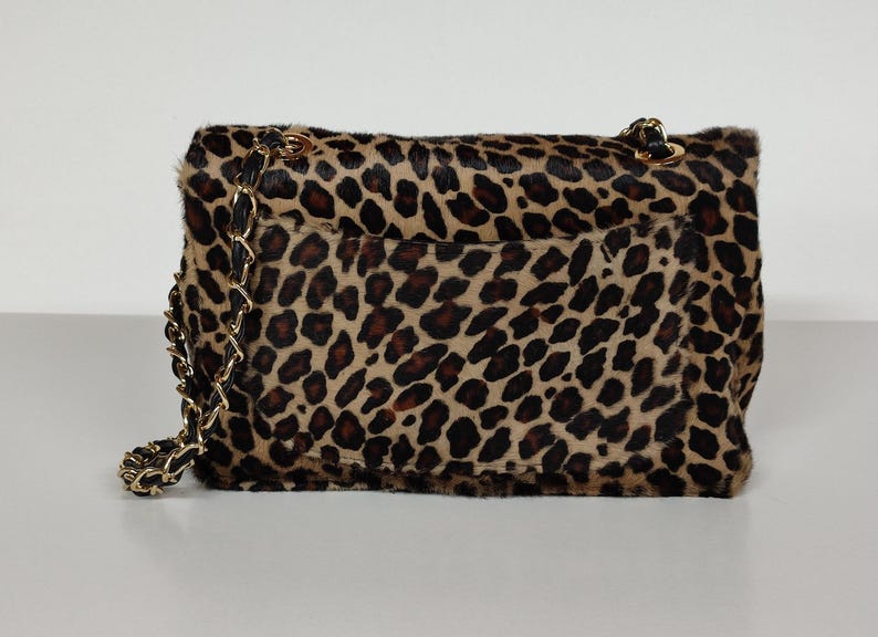 May include: A rectangular shoulder bag featuring a textured leopard print pattern in shades of tan, brown, and black. The bag includes a gold-tone metal chain strap interwoven with black material and a flap closure with a visible exterior pocket.
