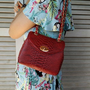 May include: A red handbag with a textured, reptile-like pattern. The bag has a gold-colored clasp and a chain strap with red leather accents. The bag is being held by a person wearing a light blue floral dress.