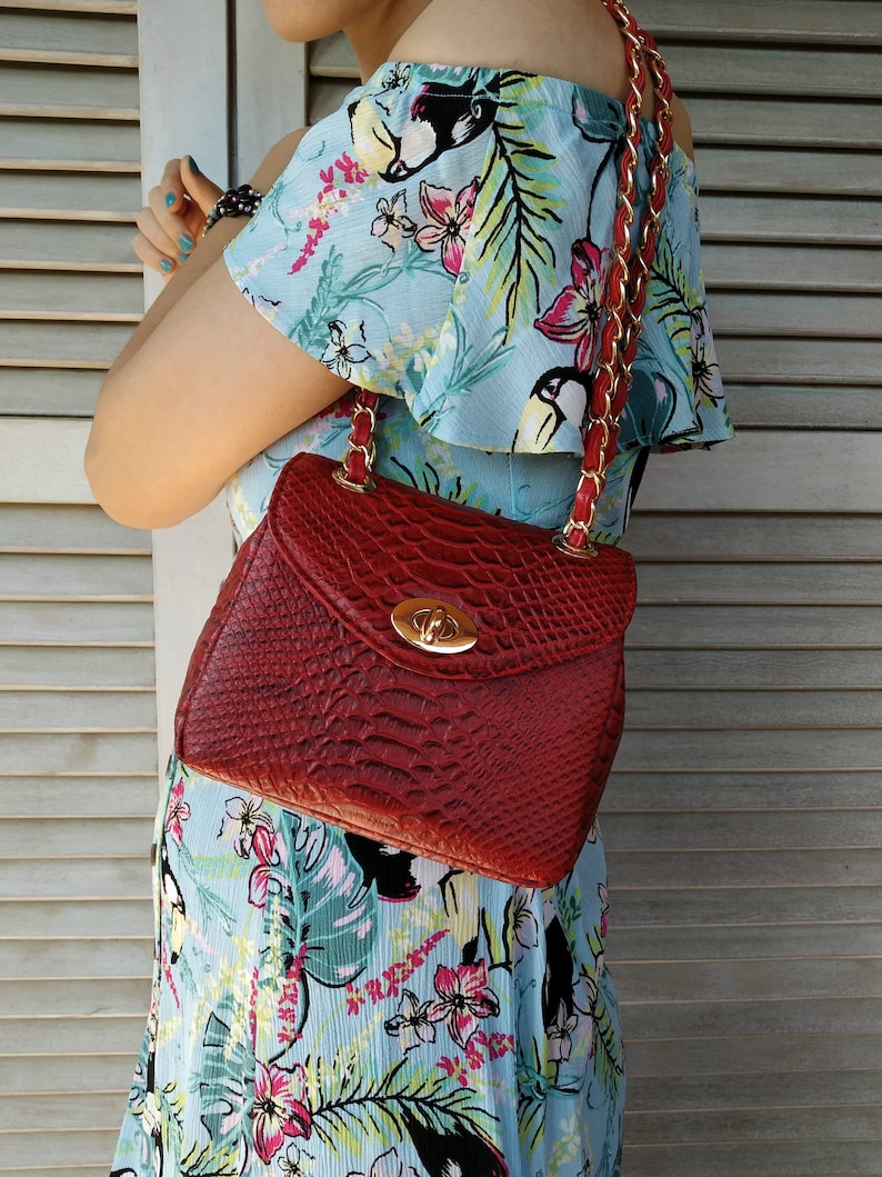 May include: A red handbag with a textured, reptile-like pattern. The bag has a gold-colored clasp and a chain strap with red leather accents. The bag is being held by a person wearing a light blue floral dress.