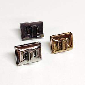 May include: Three rectangular metal twist locks for bags, featuring a smooth, reflective finish. One lock is dark gunmetal grey, one is polished silver, and one is bright gold. Each piece has a central rotating latch mechanism for secure closure.