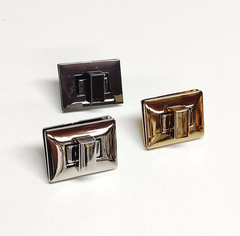 May include: Three rectangular metal twist locks for bags, featuring a smooth, reflective finish. One lock is dark gunmetal grey, one is polished silver, and one is bright gold. Each piece has a central rotating latch mechanism for secure closure.