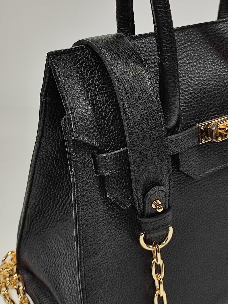 May include: Close-up of a black leather handbag with a textured finish. The bag features a gold-colored clasp and chain strap. The shoulder strap is also black leather.