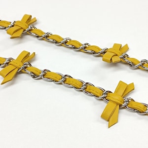 K&ouml;nnte beinhalten: Nahaufnahme von zwei gelben Lederketten mit silberfarbenen Metallgliedern. Jede Kette hat eine kleine, gebundene Schleife aus dem gleichen gelben Leder. Die Ketten sind vor einem wei&szlig;en Hintergrund angeordnet.