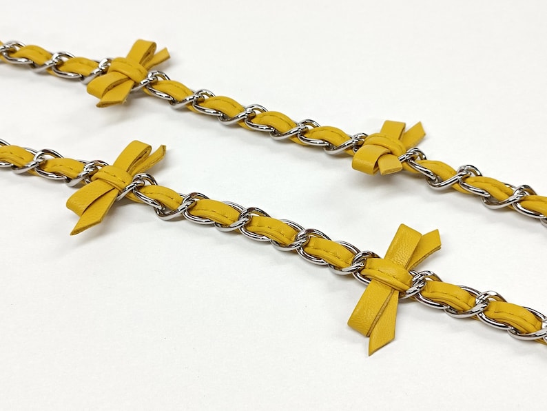 K&ouml;nnte beinhalten: Nahaufnahme von zwei gelben Lederketten mit silberfarbenen Metallgliedern. Jede Kette hat eine kleine, gebundene Schleife aus dem gleichen gelben Leder. Die Ketten sind vor einem wei&szlig;en Hintergrund angeordnet.