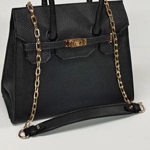 May include: Black handbag with a gold chain strap and accents. The bag has a textured surface, a flap closure, and a top handle. The gold chain is draped over the bag and can be used as a shoulder strap.