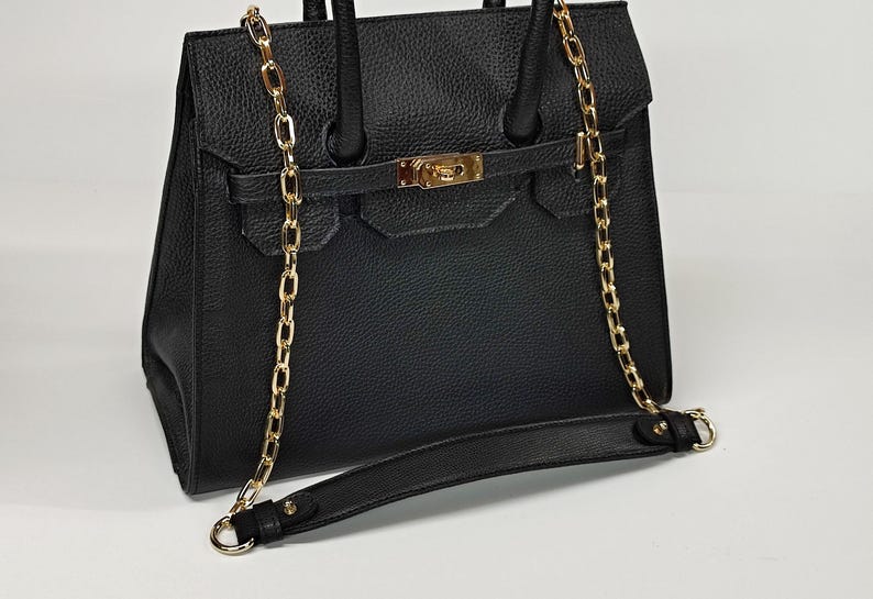 May include: Black handbag with a gold chain strap and accents. The bag has a textured surface, a flap closure, and a top handle. The gold chain is draped over the bag and can be used as a shoulder strap.