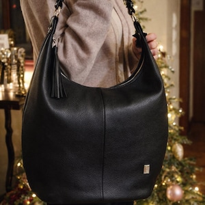 May include: A black hobo bag with a pebbled texture, featuring a single shoulder strap with metallic hardware, a decorative black tassel hanging from the side, a top zipper closure, and a small rectangular silver-toned logo plaque on the lower front.