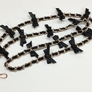 K&ouml;nnte beinhalten: Ein dekorativer Kettenriemen mit einer goldfarbenen Kette und schwarzen Lederdetails. Der Riemen ist mit kleinen schwarzen Schleifen verziert. An den Enden des Riemens befinden sich goldfarbene Verschl&uuml;sse.