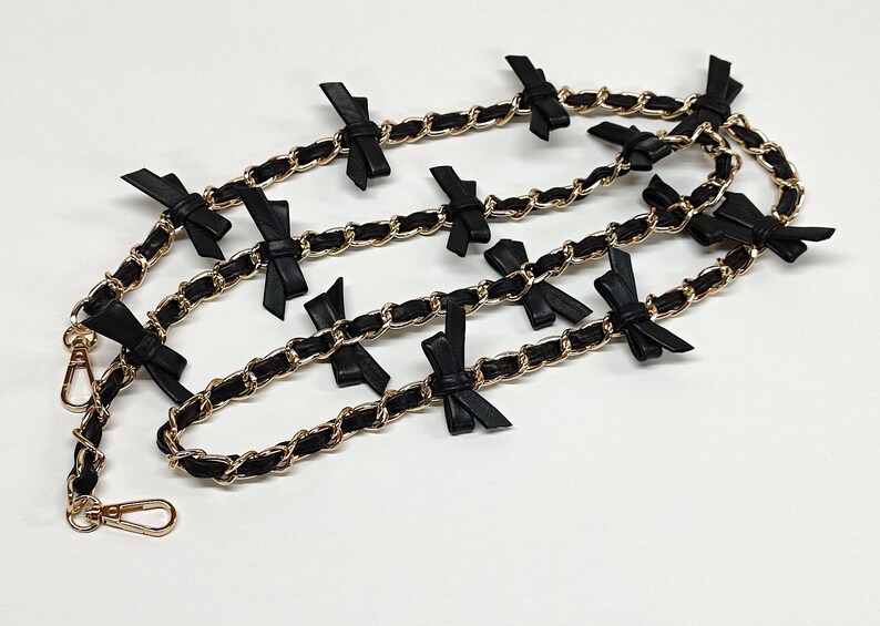 K&ouml;nnte beinhalten: Ein dekorativer Kettenriemen mit einer goldfarbenen Kette und schwarzen Lederdetails. Der Riemen ist mit kleinen schwarzen Schleifen verziert. An den Enden des Riemens befinden sich goldfarbene Verschl&uuml;sse.