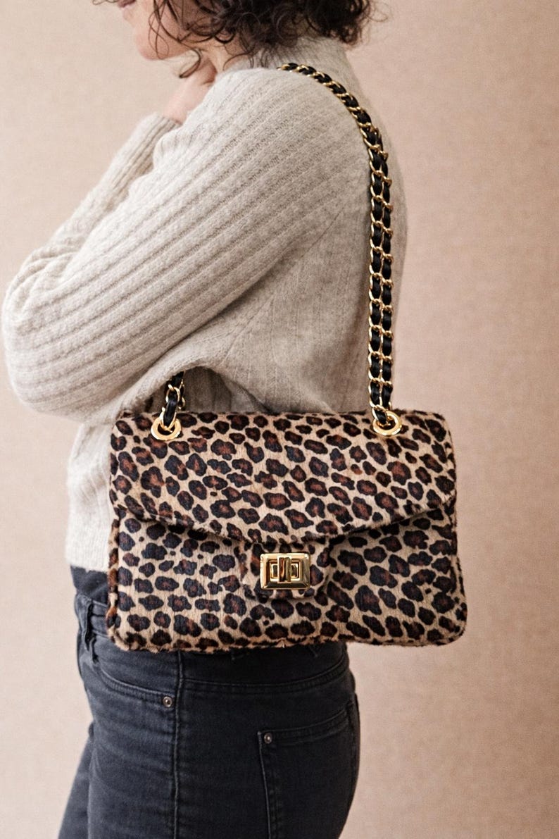 May include: A rectangular handbag with a leopard print pattern. The bag has a gold-colored clasp and a chain strap with black leather accents. The bag is being worn over the shoulder.