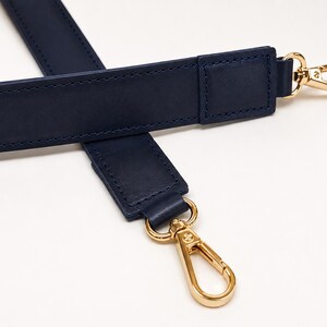 May include: A navy blue bag strap featuring smooth, matte-finish material with tonal stitching along the edges. The strap is equipped with polished gold-tone metal lobster claw clasps at both ends, allowing for secure attachment to various bags.