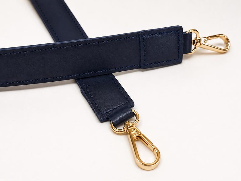 May include: A navy blue bag strap featuring smooth, matte-finish material with tonal stitching along the edges. The strap is equipped with polished gold-tone metal lobster claw clasps at both ends, allowing for secure attachment to various bags.