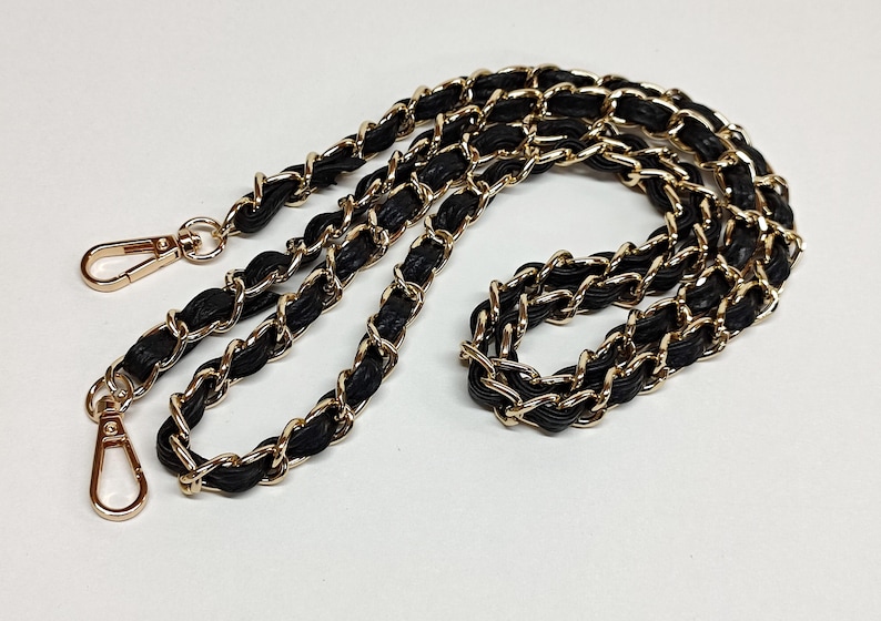 May include: Black and gold chain strap with two gold clasps. The chain is made of interwoven leather and metal.