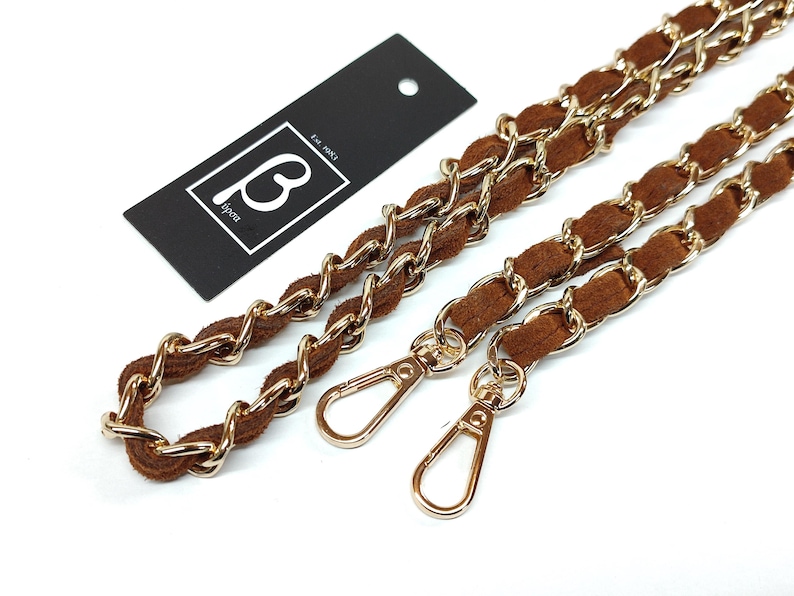 May include: A brown suede and gold-tone metal chain strap for a handbag, featuring two lobster claw clasps at the ends. A black rectangular tag is included with the white text "B" and "est. 1983" printed inside a white square border.