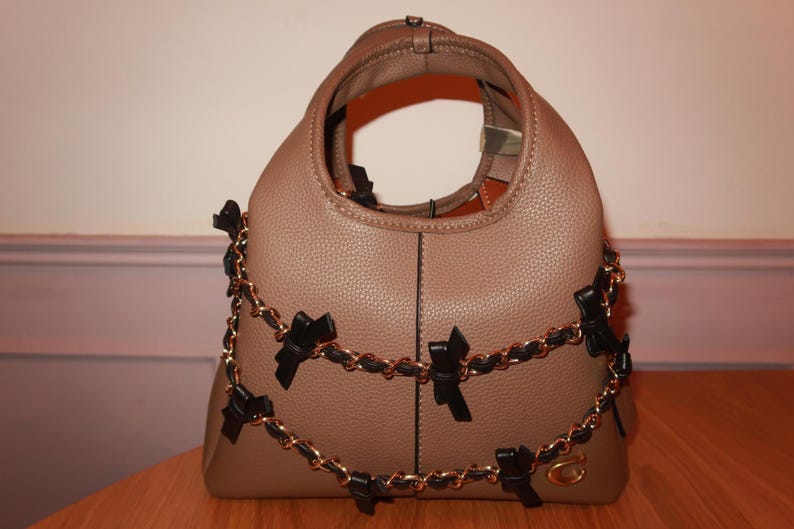 K&ouml;nnte beinhalten: Taupefarbene Handtasche mit strukturierter Oberfl&auml;che. Die Tasche hat einen goldfarbenen Kettenriemen, der mit schwarzen Schleifen verziert ist. Die Tasche hat eine runde Form und einen einzelnen Griff.