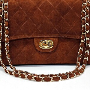 May include: A brown suede handbag featuring a quilted diamond pattern, a gold-tone metal turn-lock closure, and a long chain strap interwoven with matching brown suede material. The rectangular bag has a structured flap and polished gold hardware.